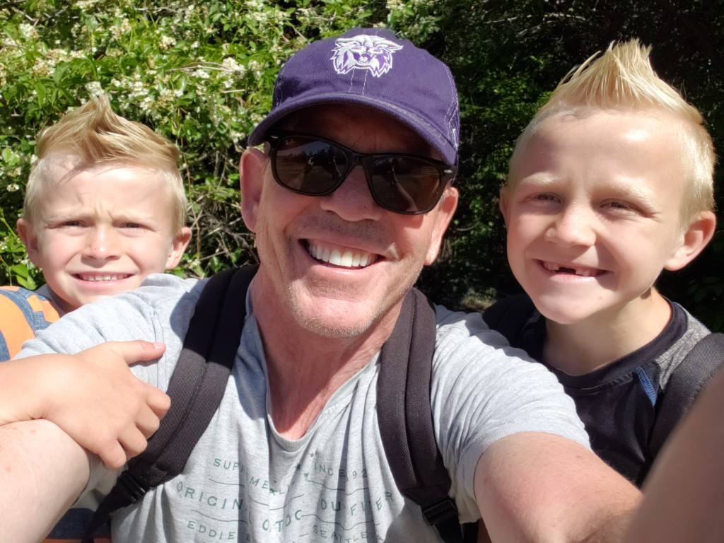 Kris Jugler smiling outdoors with two young sons in front of flowering greenery